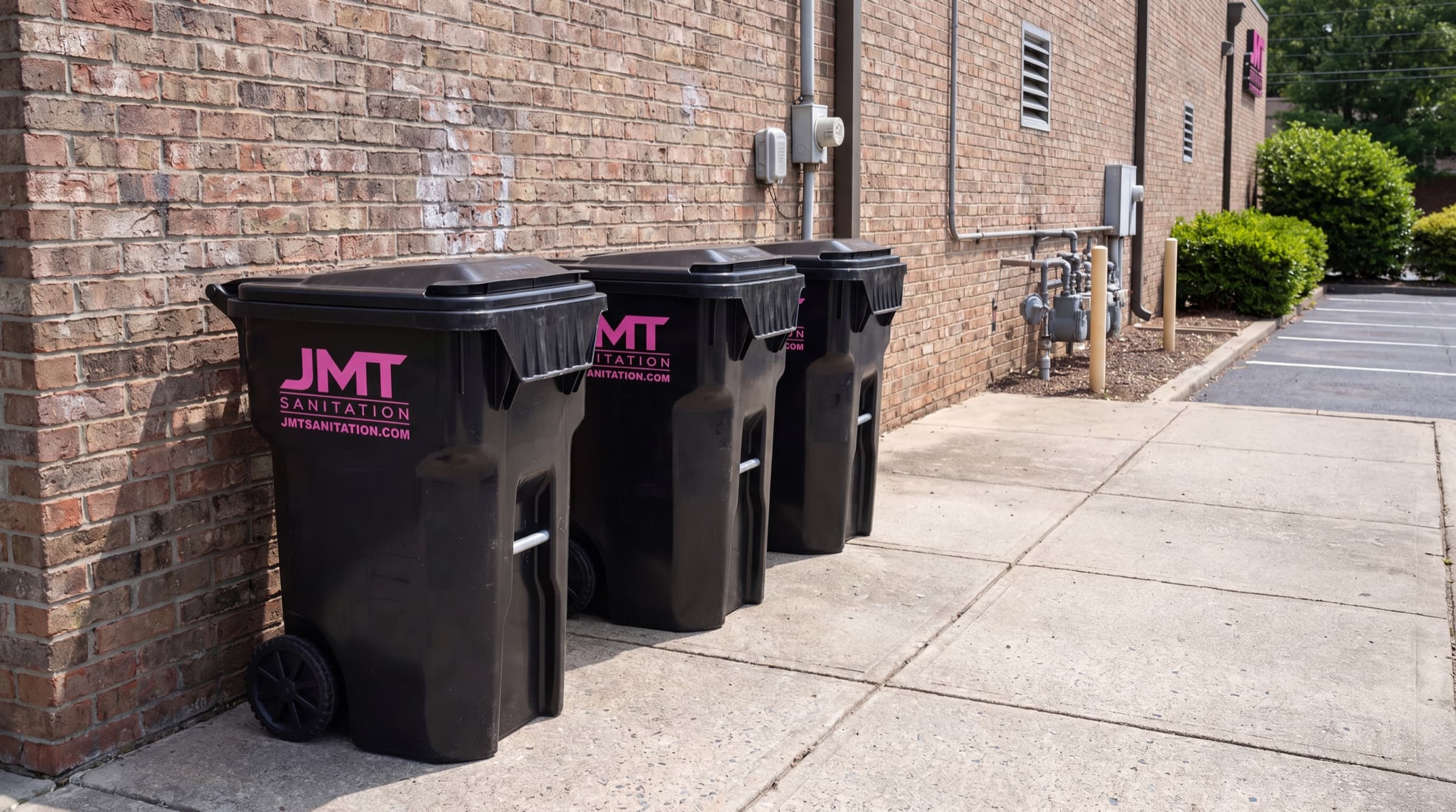 A row of three JMT Sanitation business garbage totes lined up against the brick wall of a commercial building.